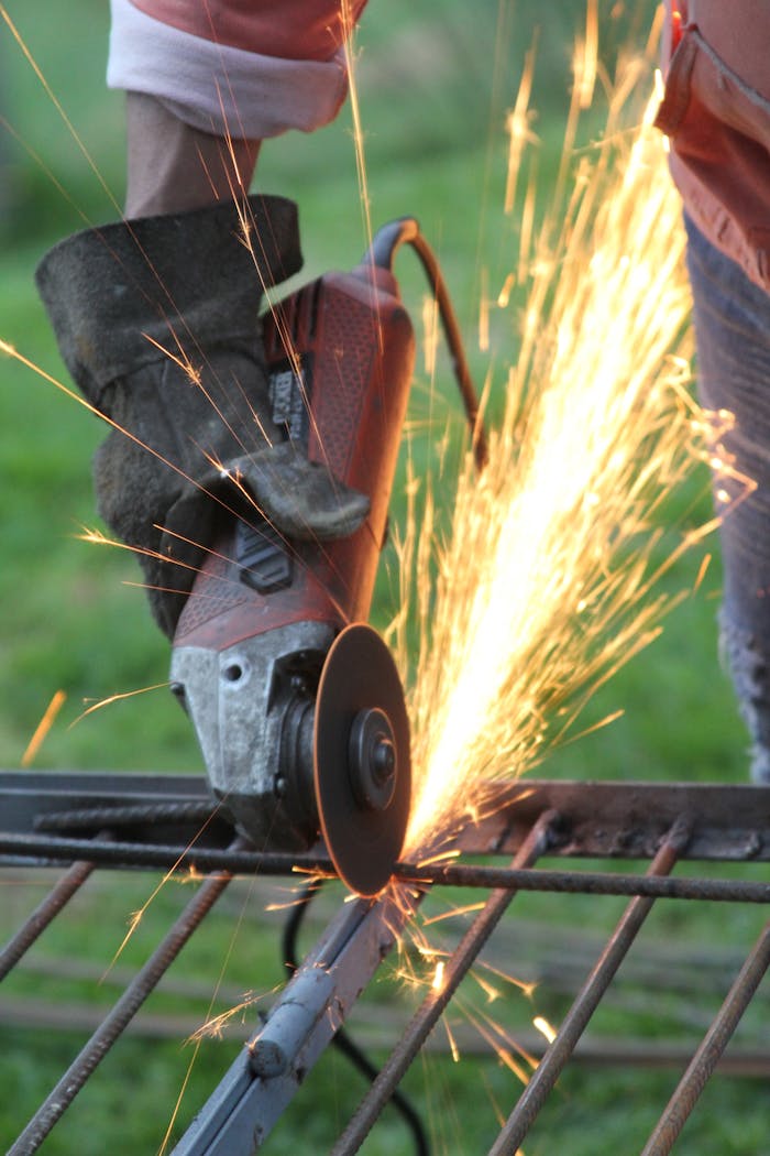 A worker wearing gloves uses an angle grinder on metal, creating sparks.