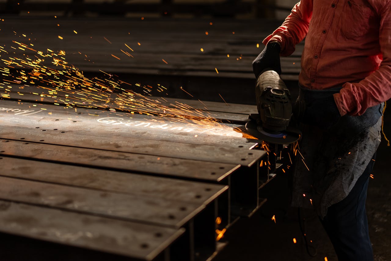 services-03 A worker grinds metal beams with an angle grinder, creating vibrant sparks in an industrial setting.